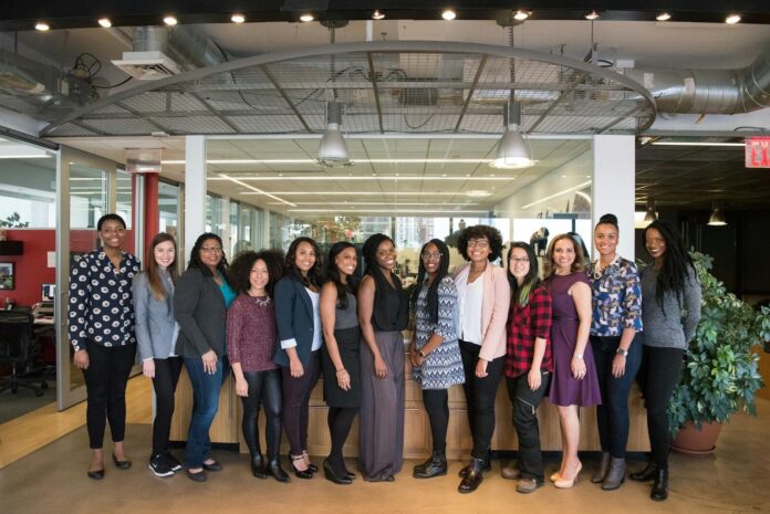 Photo by Christina Morillo A diverse group of women in technology poses together in an office lobby, exuding confidence and teamwork.