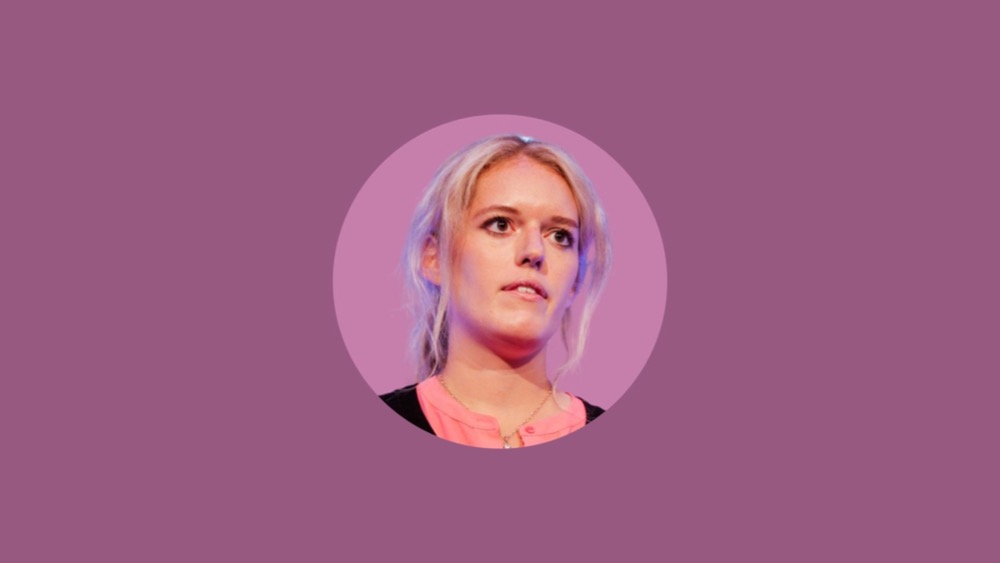 A person with blonde hair and a serious expression is shown against a purple background. They are wearing a pink top and a black hybrid jacket.