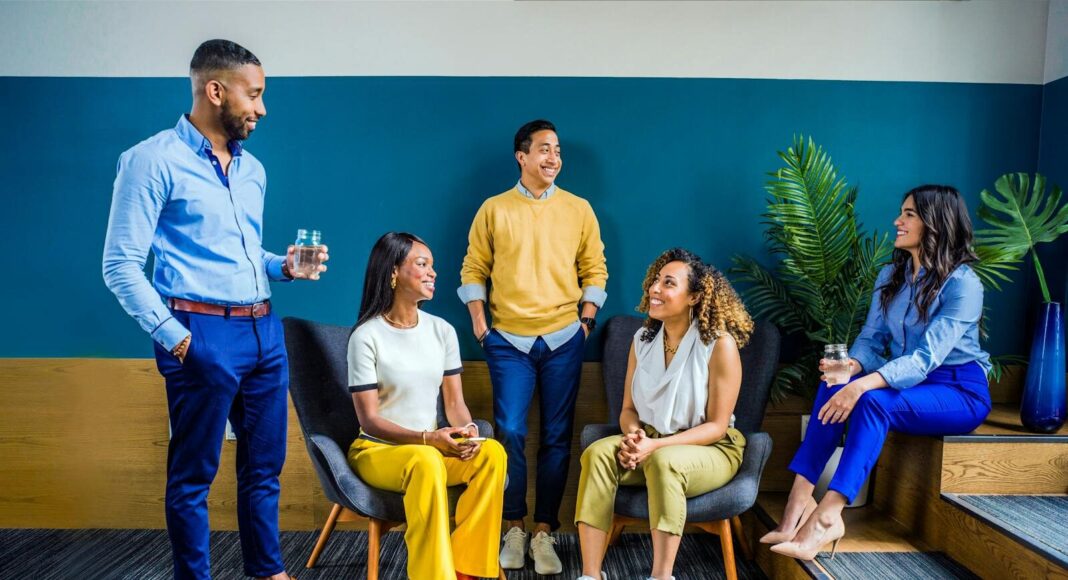 A diverse group of five professionals engaging in a casual indoor meeting, smiling and talking.