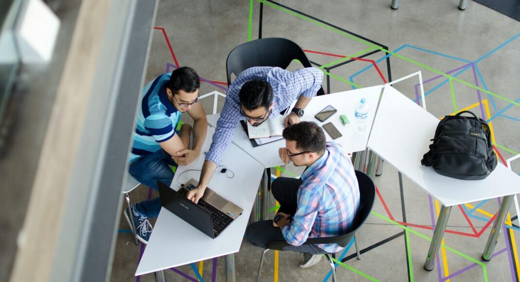 Three men collaborating over a laptop in a modern, geometric-themed office space.