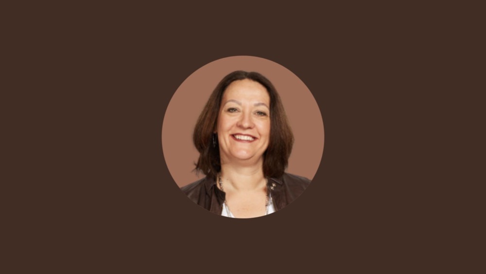 Claire Scott, a woman with shoulder-length brown hair and a big smile, stands against a solid brown background. She is wearing a dark-colored jacket, embodying the balance needed to avoid burnout amidst rising tech expectations.