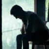 A man sits alone in a moody indoor setting, silhouetted against a bright window, deep in thought.