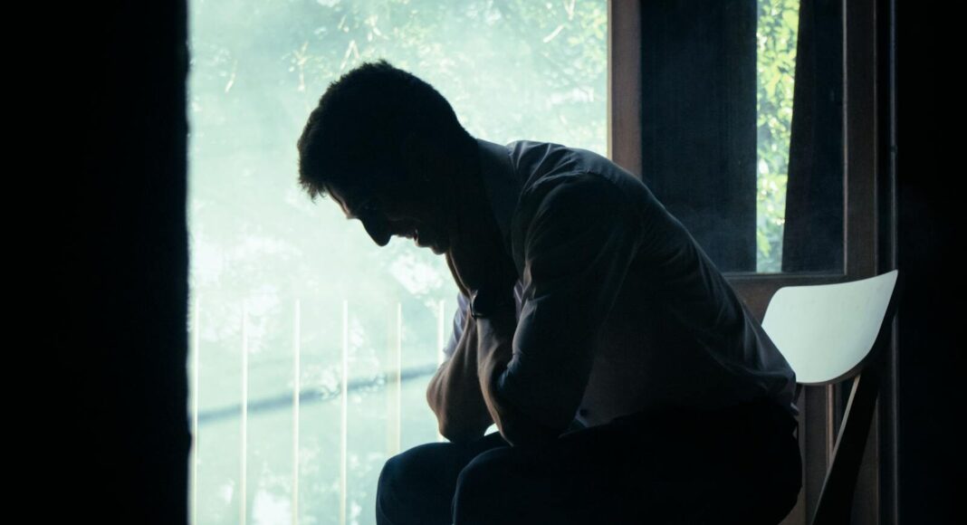 A man sits alone in a moody indoor setting, silhouetted against a bright window, deep in thought.