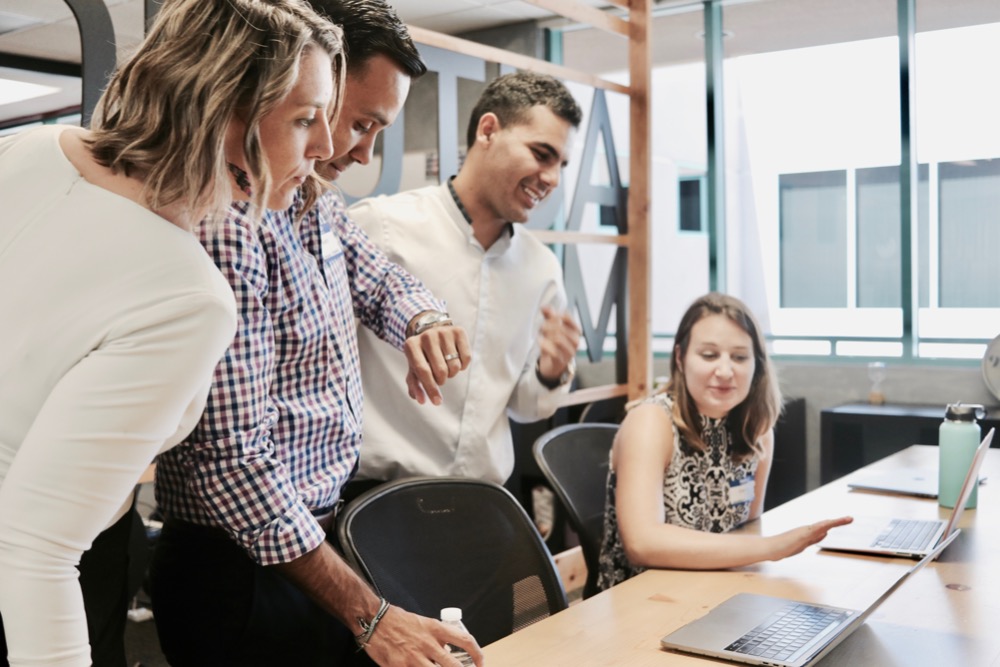 A group of four colleagues stand and sit around a desk, looking at a laptop screen. One person is gesturing with their hand while others are engaged and smiling, perhaps discussing the benefits of their new four-day week.