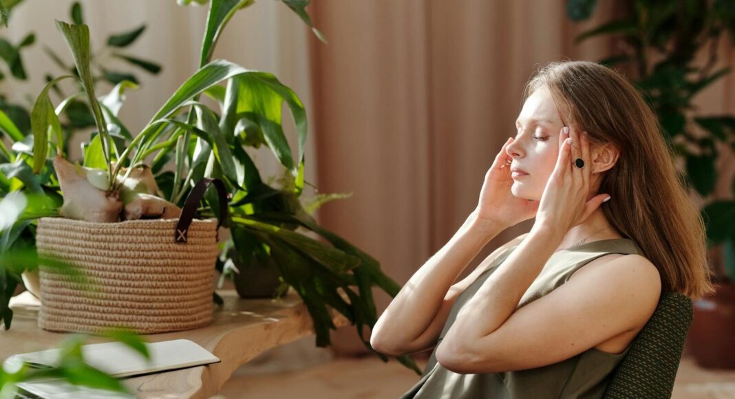 A woman relaxes, massaging her temples among indoor plants in a bright, sunlit room.