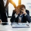 An overwhelmed businessman grasps his head, surrounded by reports in a bright office.