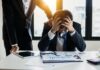 Millions of workers affected by ‘secondhand stress’ from colleagues An overwhelmed businessman grasps his head, surrounded by reports in a bright office.