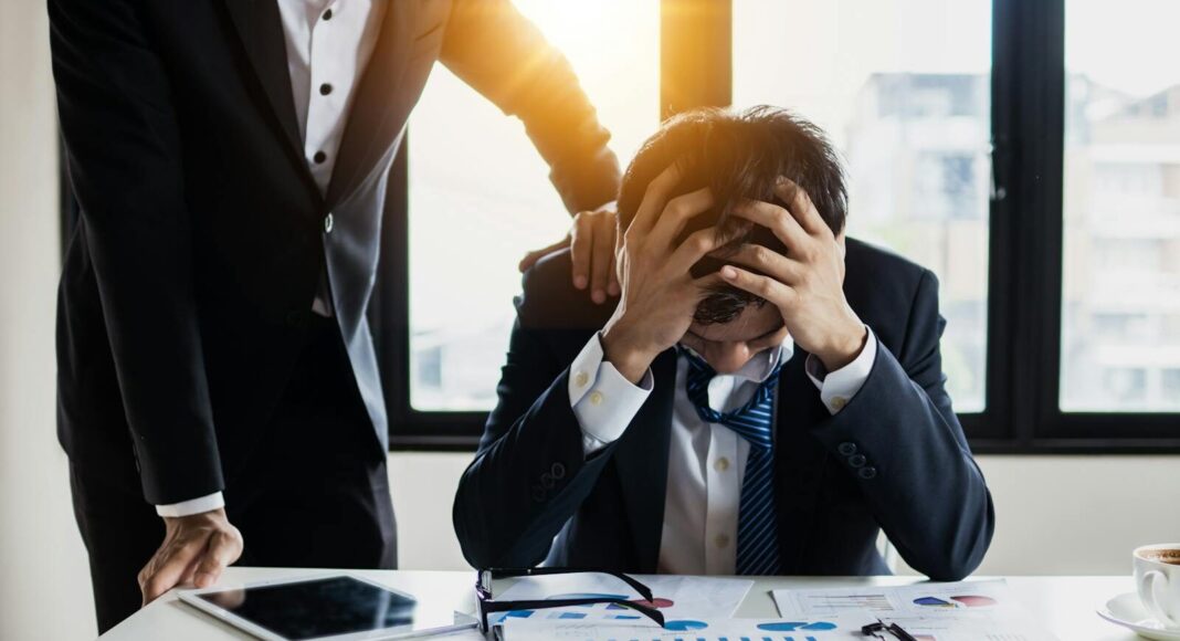 An overwhelmed businessman grasps his head, surrounded by reports in a bright office.