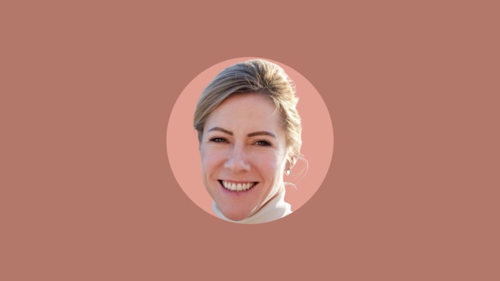 A woman with a light complexion smiles at the camera against a solid pinkish-brown background. She has blonde hair pulled back and wears a white top, her expression radiating positive feedback.