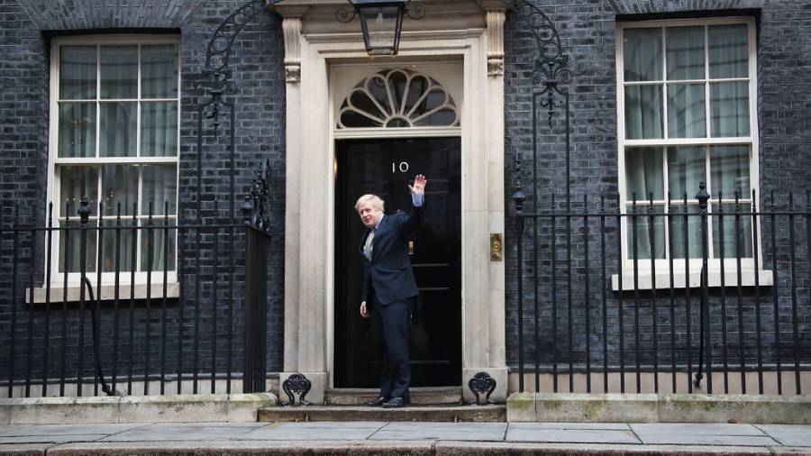 A man in a suit waves while standing in front of the black door marked with the number 10 at an official-looking building with black bricks and white trim, perhaps reflecting his role in personnel management.
