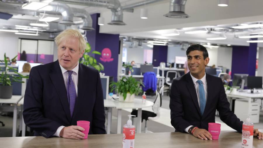 Two men, one with blond hair and the other with dark hair, are standing in an office holding pink cups. Casually dressed in suits, they are surrounded by desks, computers, plants, and office decor—an everyday scene of personnel management at work.