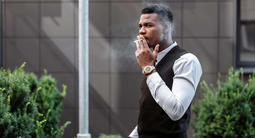 A stylish young man smoking a cigarette outside an urban building.