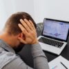 A man with his hands on his head, indicating stress and worries, sits in front of a desk with a laptop.