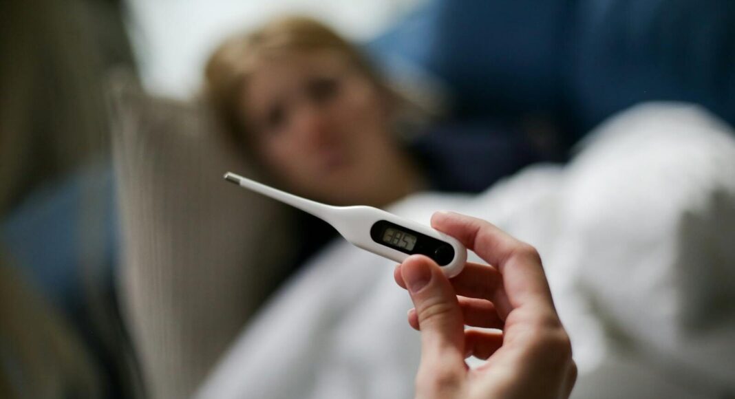 Hand holding a digital thermometer with blurred background of a sick person.