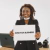 A smiling woman with dreadlocks holding an 'Open for Business' sign in an indoor setting.