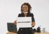 Employees increasingly building businesses around their day jobs A smiling woman with dreadlocks holding an 'Open for Business' sign in an indoor setting.