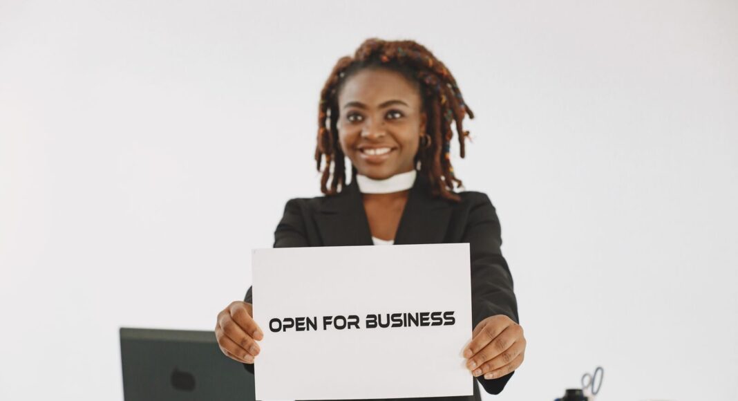 A smiling woman with dreadlocks holding an 'Open for Business' sign in an indoor setting.