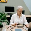 An older woman multitasking with lunch and work on laptop in an office setting.