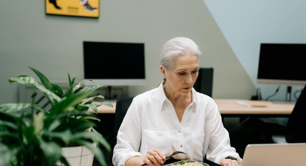 Photo by cottonbro studio An older woman multitasking with lunch and work on laptop in an office setting.