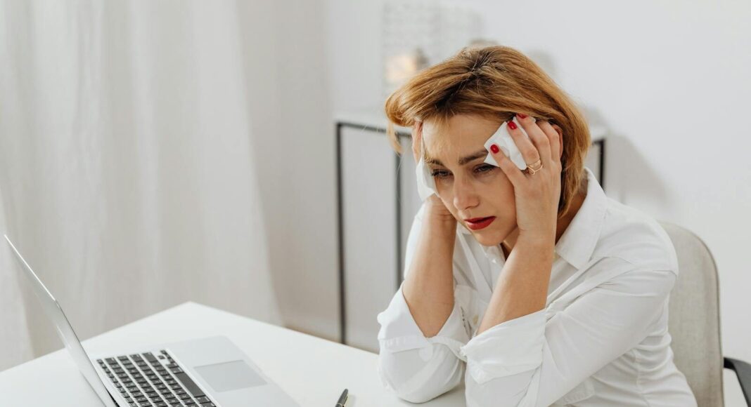 A woman in distress at her office desk, surrounded by tissues and a laptop.