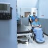 A nurse wheels a patient in a wheelchair down a hospital corridor, reflecting care and medical professionalism.