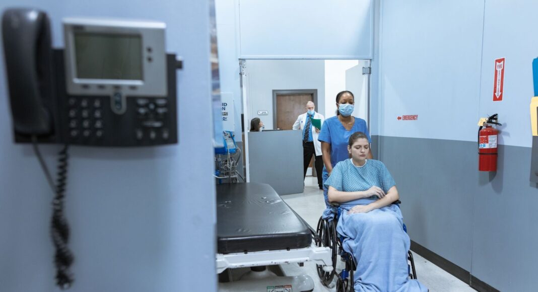 A nurse wheels a patient in a wheelchair down a hospital corridor, reflecting care and medical professionalism.