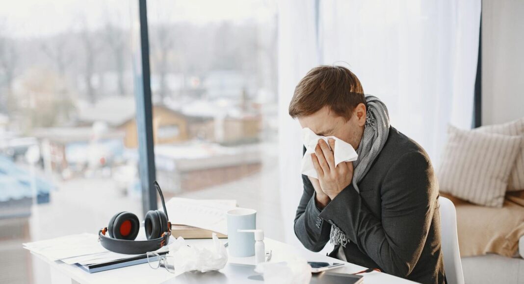 Man sneezing at home desk, indicating remote work during illness.