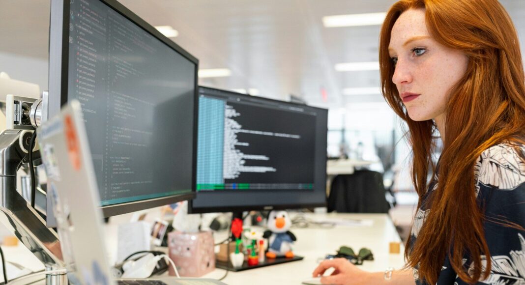 A young woman works at a desk with two monitors and a laptop.