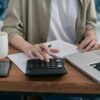 A person using a calculator and laptop at a wooden desk.