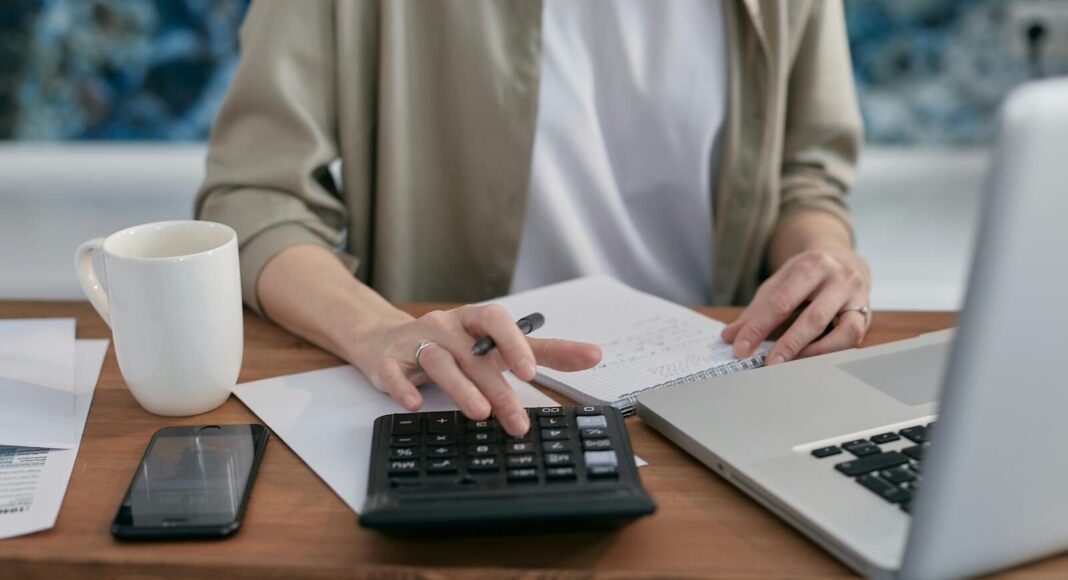A person using a calculator and laptop at a wooden desk.