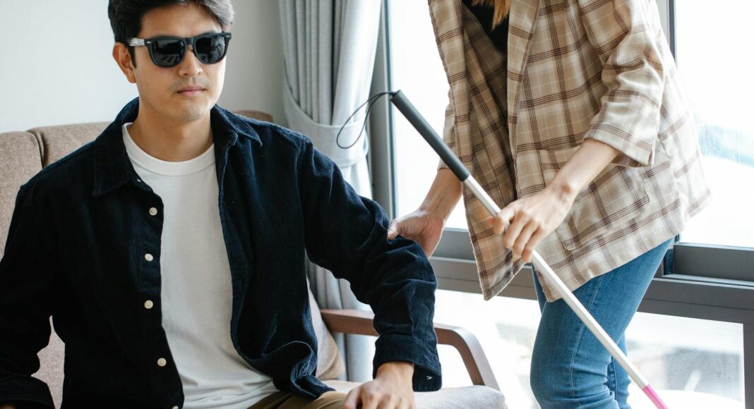 A visually impaired man receives support from a caregiver indoors, using a walking stick.