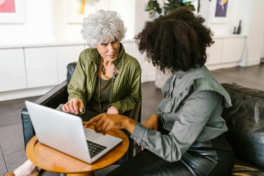 An older and a younger woman discussing work on a laptop in a modern office setting.
