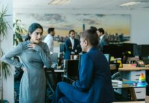 Workplace sexism still widespread, survey finds Two women talking in an office with other people behind.