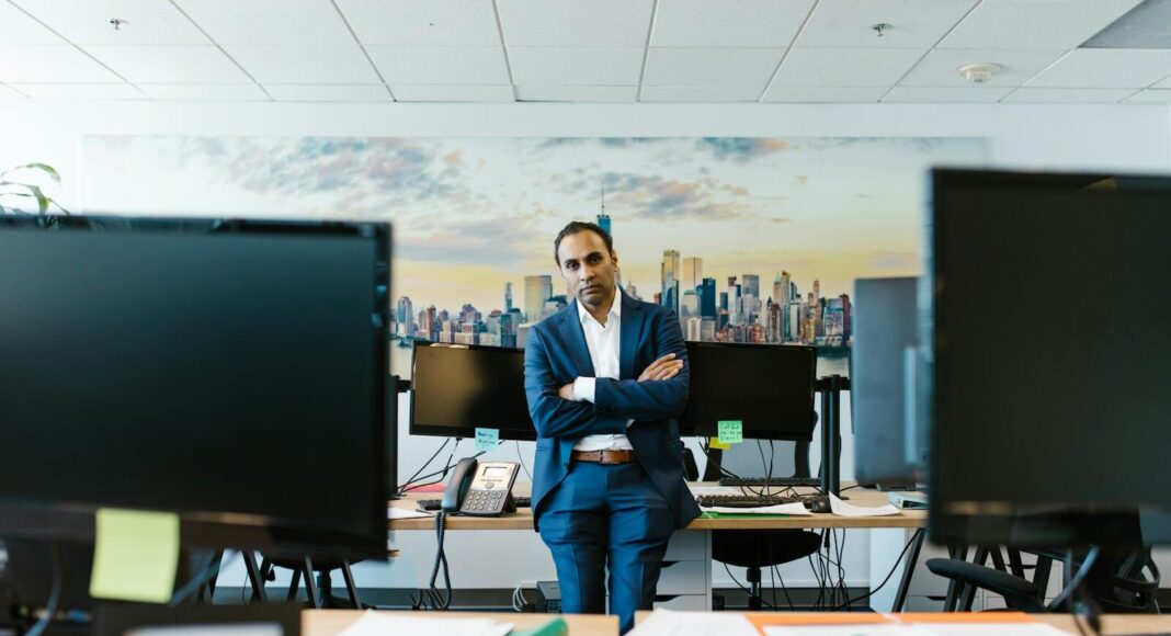 A businessman in a suit stands confidently in a modern office setting with crossed arms.