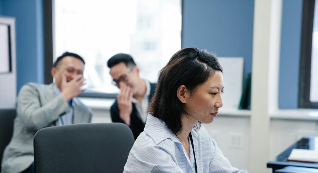 Focused office worker with colleagues gossiping in the background, showcasing workplace dynamics.