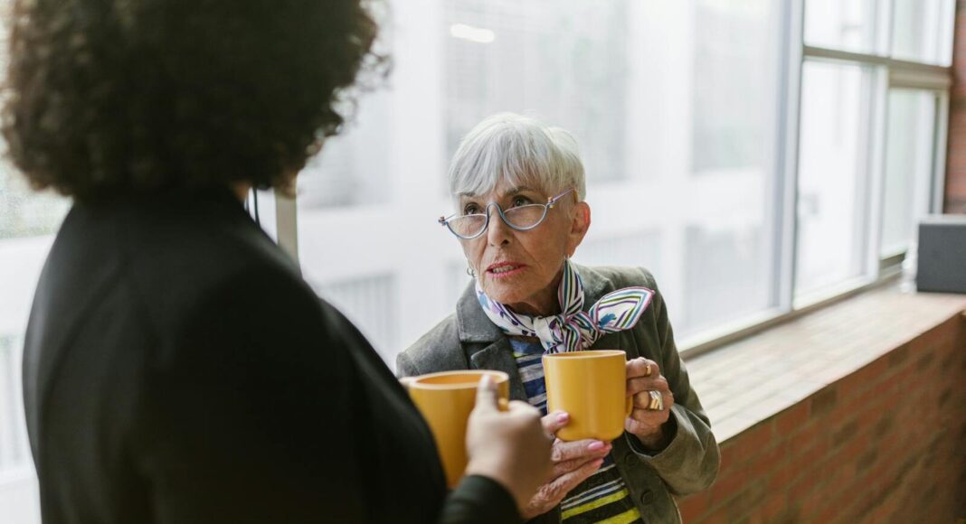 An older woman conversing with a colleague while holding coffee mugs indoors.