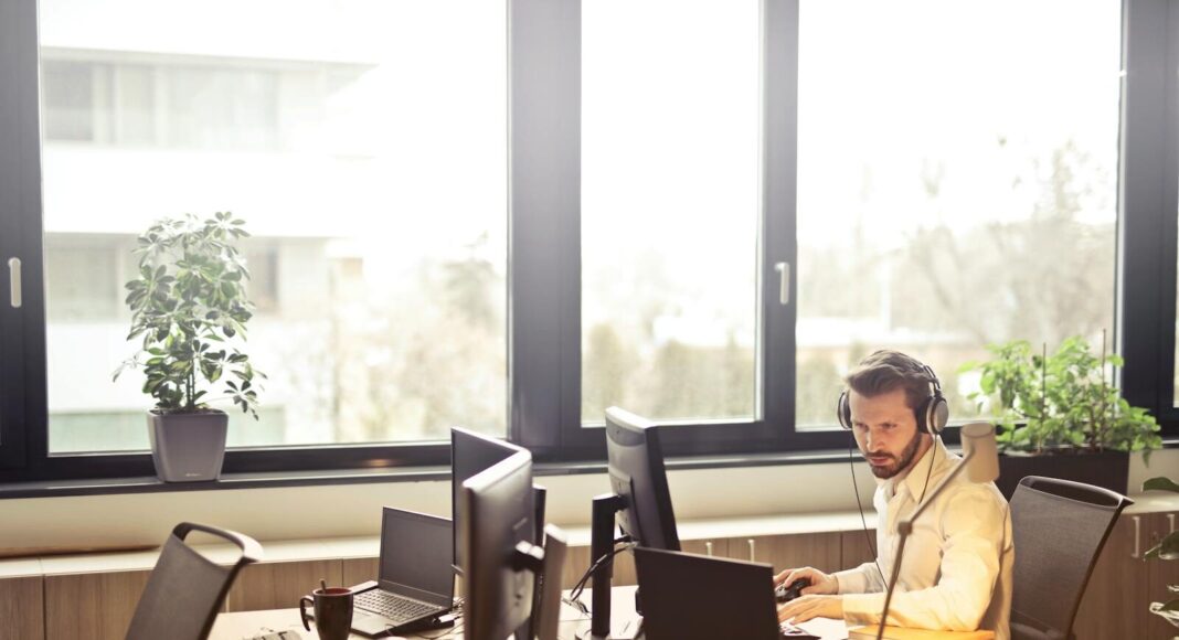 A man sits at a desk using multiple computers and a headset in a well-lit modern office.