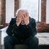 An elderly man sitting on a bed indoors, expressing regret with his head in his hands.