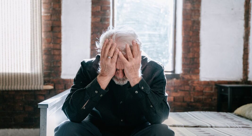 Photo by Ivan S An elderly man sitting on a bed indoors, expressing regret with his head in his hands.