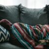 A person lying on gray sofa, wrapped in a blanket.