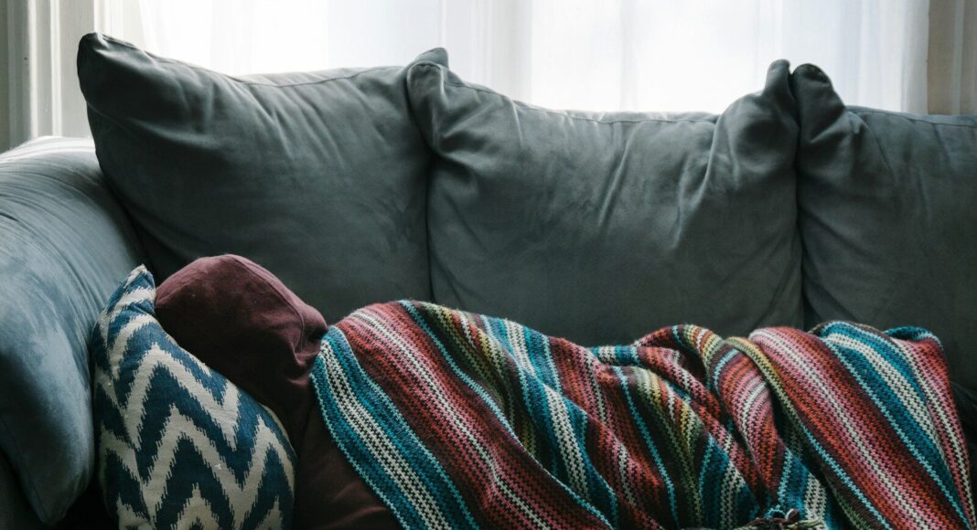 A person lying on gray sofa, wrapped in a blanket.