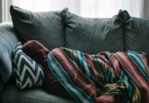Government to replace some GP sick notes with return-to-work plans A person lying on gray sofa, wrapped in a blanket.