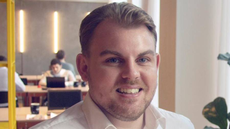 A man with short hair and a beard smiles at the camera. He wears a white shirt. The background shows HR personnel working at desks in a modern office space.