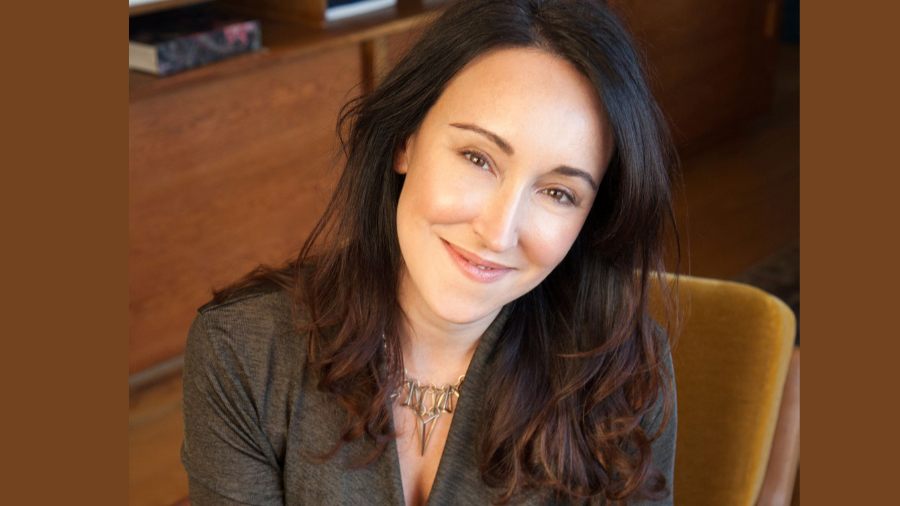 A woman with long, dark hair wearing a gray top and a necklace smiles while seated indoors with a wooden cabinet in the background, seemingly free from any email apnoea stress.