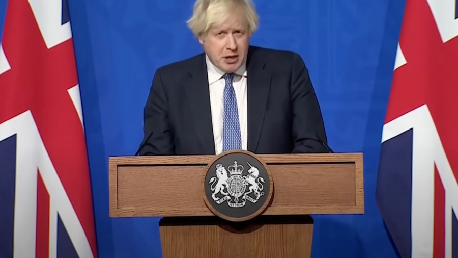 A person with light hair stands at a podium with a crest, flanked by two Union Jack flags, discussing Plan B measures in response to the Omicron variant against a blue background.