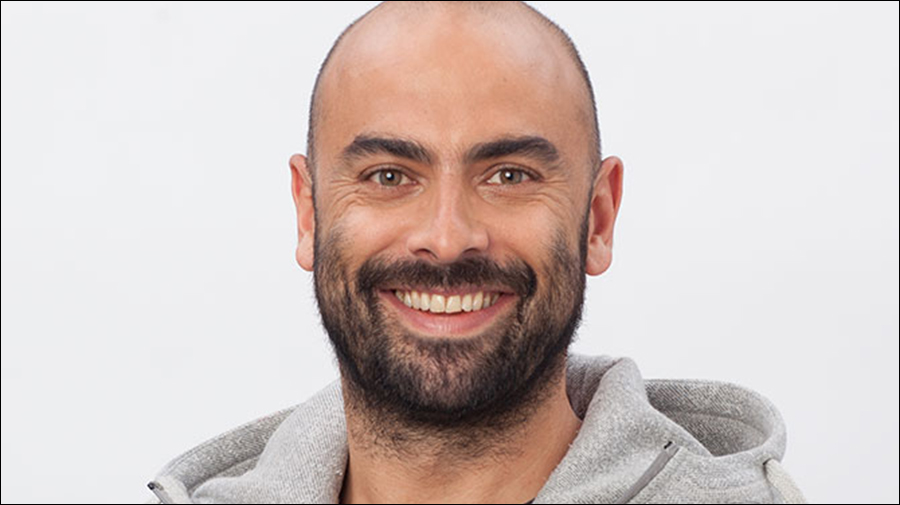 A bald man with a beard smiles while wearing a light gray hoodie against a plain white background, embodying the approachable and personable qualities essential in Human Resources.