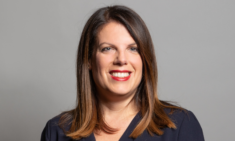 A woman with shoulder-length brown hair and a navy blue top, representing women in work, smiles against a gray background.