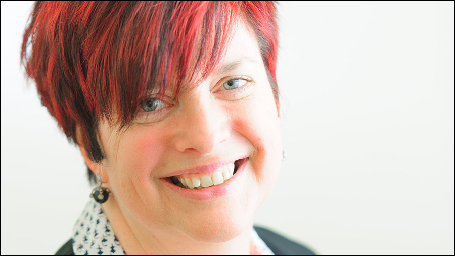 A person with short red hair and a friendly smile in a well-lit room, wearing a white and black patterned shirt and a pair of earrings, exudes professionalism expected in HR roles.
