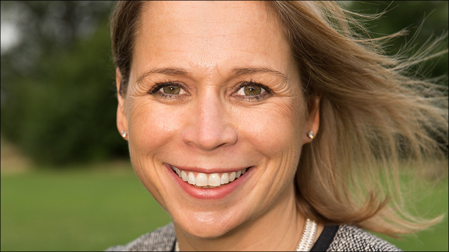 A woman with blonde hair and a smile on her face is looking directly at the camera. She is outdoors with a green background, exuding confidence that comes from her expertise in people management.