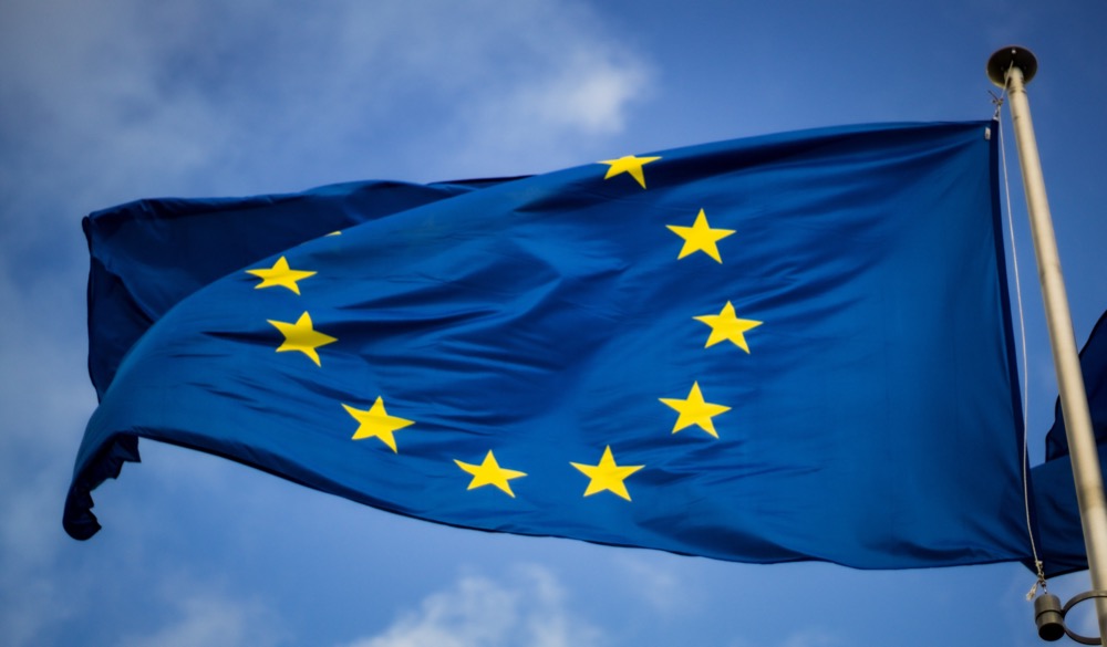A blue flag with twelve yellow stars in a circle, representing the European Union, flutters in the wind against a blue sky with some clouds. This symbolic representation underscores the unity and cooperation that EU recruitment strives to bring across its member countries.